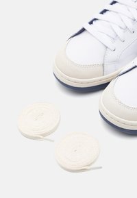 Vita sneakers med beige suéde detaljer, rund marinblå sula och platta vita skosnören. Snörena visas ihopvirade bredvid skorna.