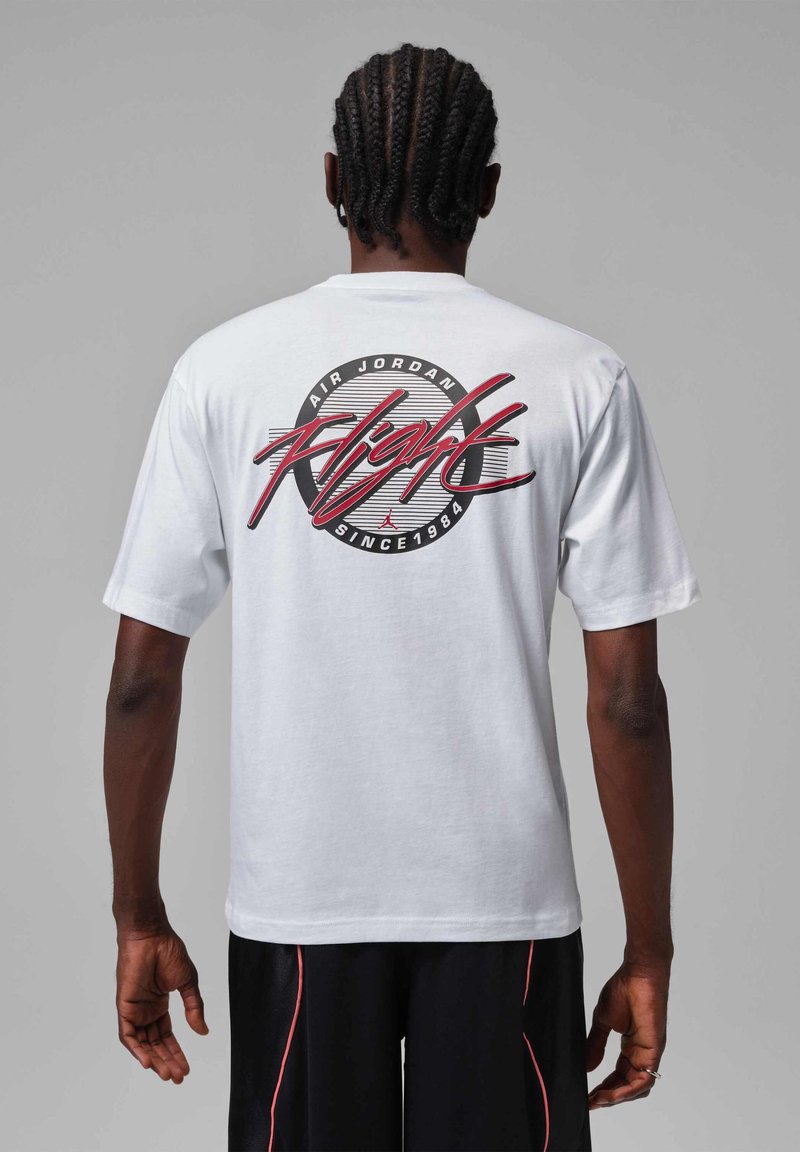 Hombre con cabello trenzado, vistiendo una camiseta blanca Air Jordan Flight y pantalones cortos negros con rayas rojas, de pie sobre un fondo gris.