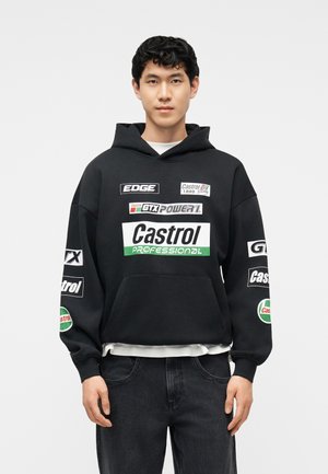 Young man wearing a black hoodie with Castrol and racing logos, standing against a plain light background.