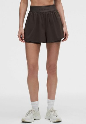 Mujer con pantalones cortos deportivos negros de Lululemon, calcetines blancos y zapatillas blancas para correr, de pie frente a un fondo claro y liso.
