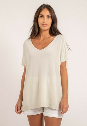 Femme aux longs cheveux noirs portant un haut ample à manches courtes écru et un short blanc retroussé, debout devant un fond uni.