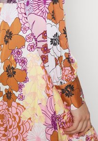 Blomsterkjole med store orange og lilla blomster på en pastel baggrund. Lange mønstrede ærmer med en tætsiddende manchet og knapper.