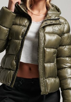 Femme portant une doudoune brillante vert olive ouverte par-dessus un débardeur blanc court et un pantalon noir, montrant un collier en chaîne dorée.