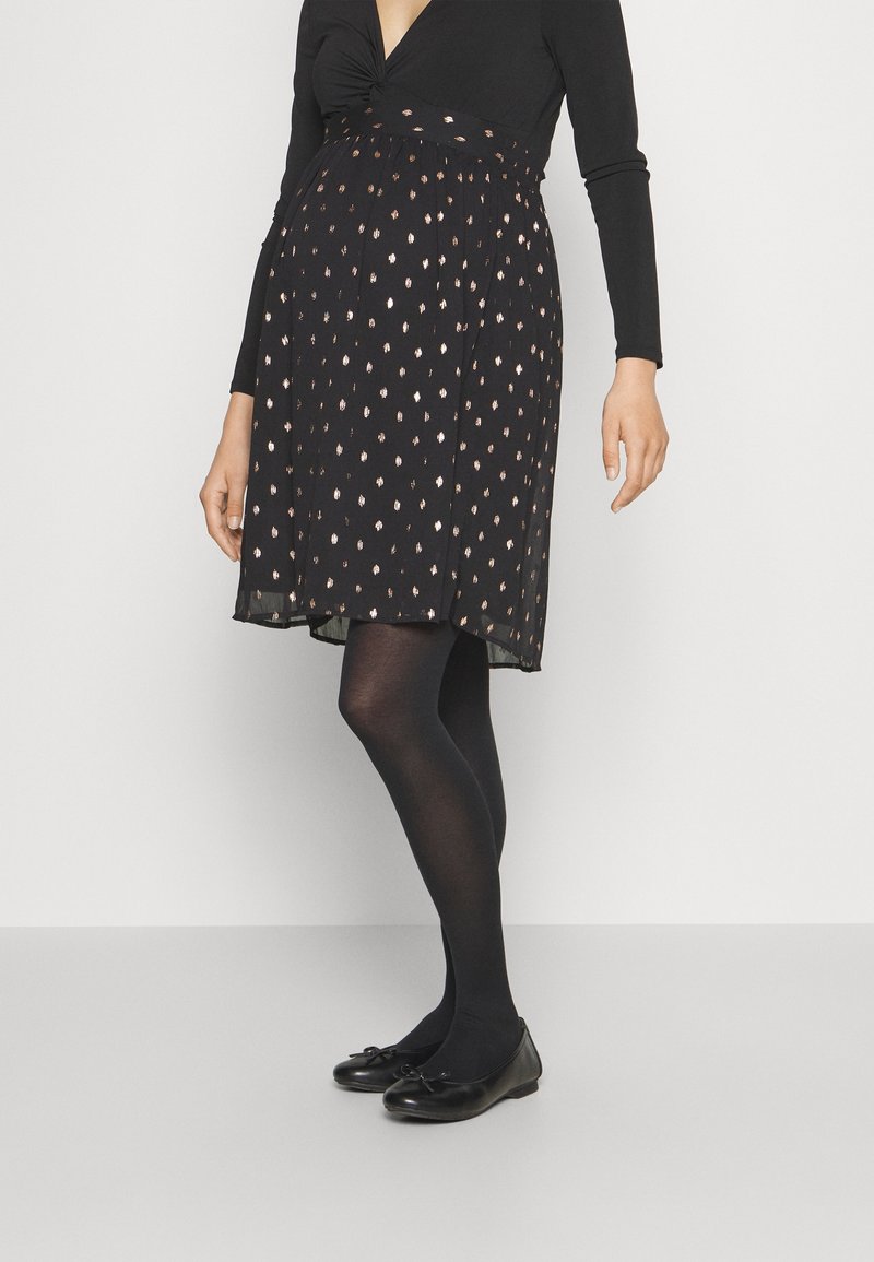 Femme portant un haut noir à manches longues, une jupe noire à pois métalliques, des collants noirs et des ballerines noires, sur un fond clair uni.