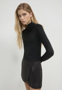 Giovane donna con lunghi capelli biondi che indossa un top nero a maniche lunghe con dettagli a volant sul collo e una gonna nera a portafoglio su sfondo bianco.