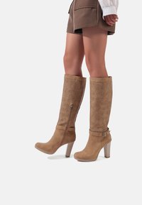 Stivali marroni alti fino al ginocchio con un design perforato, dotati di una zip laterale e un tacco grosso, realizzati in materiale morbido simile alla suede.