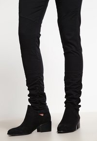 Svarta ankelskängor med mocka-textur, en låg design och en staplad klack, kombinerat med åtsittande svarta byxor med rynkdetalj.