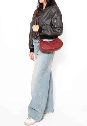 Sac à main en cuir rouge avec une surface texturée, une forme arrondie et une bandoulière ajustable. Porté avec une veste bomber noire et un jean bleu clair.
