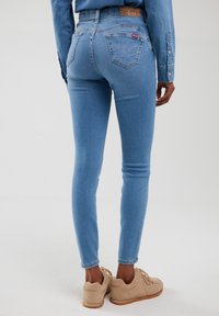 Pogled s leđa na osobu koja nosi svijetloplave uske traperice, odgovarajuću traper košulju i bež tenisice, stojeći na jednobojnoj svijetloj pozadini.