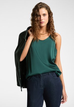 Grüne Satin-Camisole mit lockerer Passform, kombiniert mit dunklen Jeans. Ein schwarzer Mantel ist lässig über eine Schulter gelegt und verfügt über silberne Beschläge.