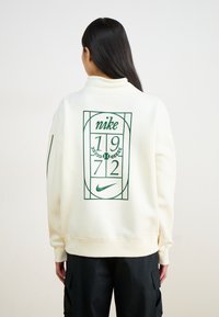 Krēmkrāsas džemperis ar zaļu grafisko dizainu, kurā redzami vārdi "nike", "1972" un tenisa motīvs uz aizmugures. Brīva piegriezuma, ribinātas piedurknes.