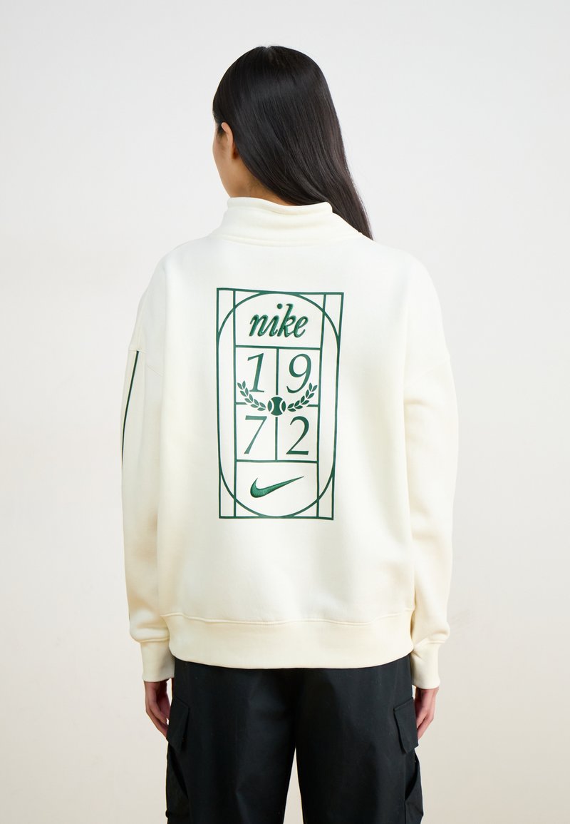 Krēmkrāsas džemperis ar zaļu grafisko dizainu, kurā redzami vārdi "nike", "1972" un tenisa motīvs uz aizmugures. Brīva piegriezuma, ribinātas piedurknes.