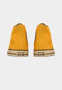 Ζευγάρι κίτρινων ψηλοτάκουνων παπουτσιών Converse All Star, εμφανιζόμενα από πίσω, σε απλό λευκό φόντο.