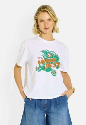 Mujer con cabello rubio corto que lleva una camiseta blanca con estampado de cítricos verdes y naranjas y pantalones cortos vaqueros azules, manos en los bolsillos.