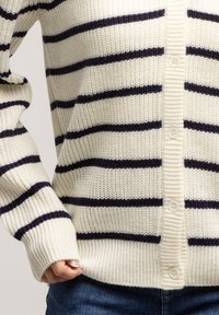 Rozpinany kardigan w kolorze kremowym z granatowymi paskami, o prążkowanej fakturze, długich rękawach i prostym wykończeniu. Posiada okrągłe guziki wzdłuż przodu.