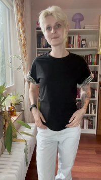 Person mit kurzen blonden Haaren, die ein schwarzes Shirt mit perlenverzierten Ärmeln und weiße Hosen trägt, steht in einem Raum mit Bücherregalen und Pflanzen.
