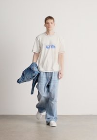 Camiseta branca com logótipo azul "HUGO", jeans largos azul claro e uma jaqueta de ganga pousada no braço, combinados com ténis brancos.