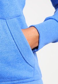 Felpa blu con polsini a costine e tasca frontale. Il tessuto texture è morbido, con cuciture a contrasto lungo le cuciture.