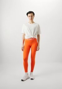 Nike orange leggings med hvidt logo på linningen, kombineret med en hvid T-shirt med knude og hvide sportssko. Glat, tætsiddende materiale.