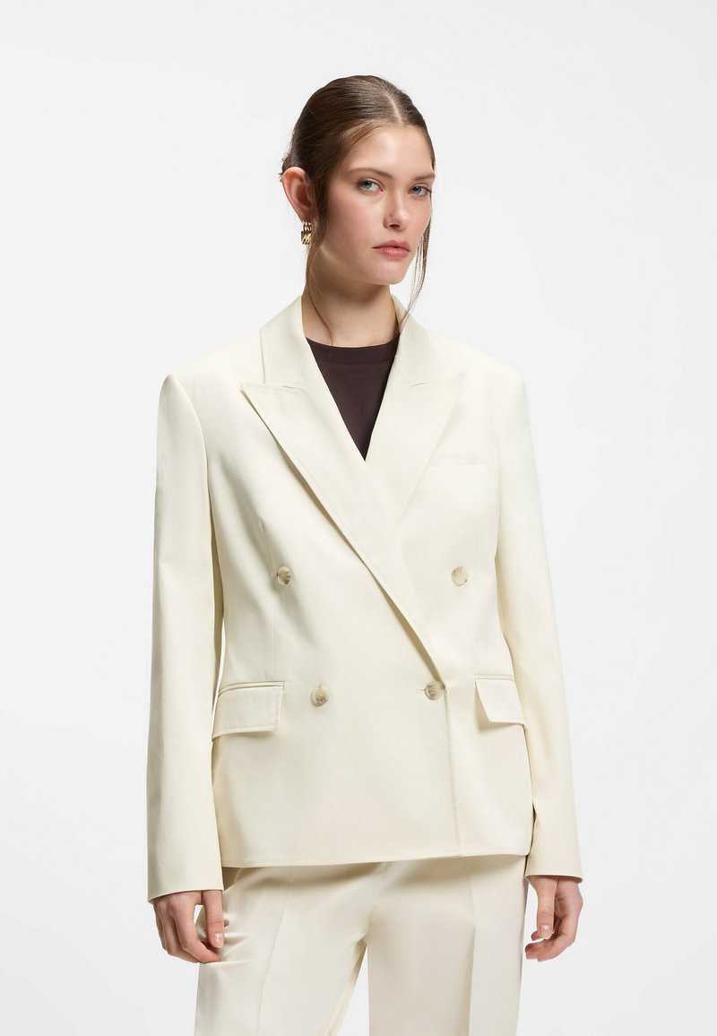 Femme aux cheveux bruns tirés en arrière, portant un blazer croisé crème et un pantalon assorti sur un haut noir, debout devant un fond blanc.