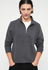 Schwarzgrauer Reißverschluss-Hoodie mit hohem Kragen, besticktem "Originals Studio"-Design auf der Vorderseite und gerippten Bündchen.