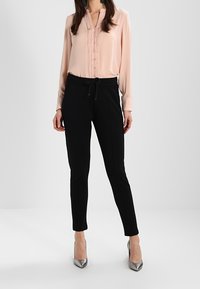 Blouse in soft pink with textured front detail, paired with black tapered pants and silver high heels. Smooth fabric and fitted design.