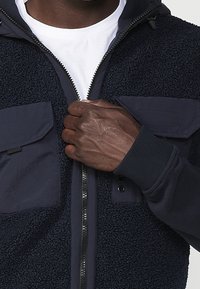 Mörkblå fleecejacka med dragkedja fram, strukturerad tyg, två bröstfickor och ribbade muddar. Vit t-shirt synlig under.