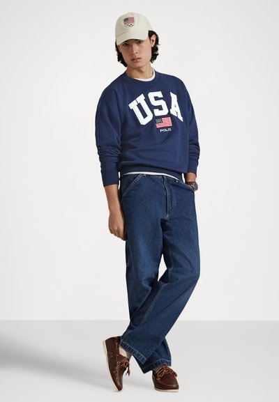 Σκούρα μπλούζα με την επιγραφή «USA» και σήμα με σημαία, συνδυασμένη με άνετο μπλε τζιν και καφέ παπούτσια. Συμπληρώνεται με εκρού καπέλο που φέρει το λογότυπο των Ολυμπιακών Αγώνων.