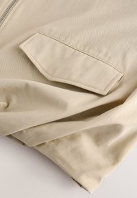 Beige stof met een gladde, matte textuur, met een gevouwen ontwerp en een rechthoekige klepzak. De hardware omvat een zilveren ritssluiting.