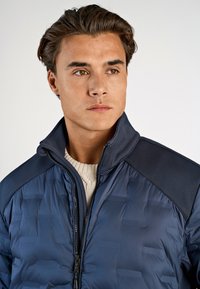 Vadderad marinblå jacka med dragkedja fram, hög krage och texturerade axeldetaljer. Bärs över en ljusfärgad tröja.