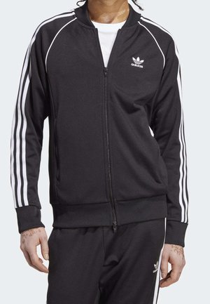 Zwarte adidas trainingsjack met ritssluiting, met witte strepen langs de schouders en mouwen, gemaakt van een gladde, gestructureerde stof.