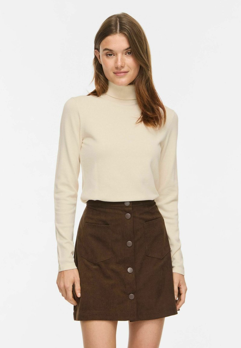 Femme portant un pull à col roulé beige à manches longues et une jupe marron avec boutons devant et poches avant, se tenant devant un fond uni.