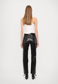 Calças de couro preto com cintura alta, costura distinta e dois bolsos traseiros com padrões abstratos. Usadas com uma t-shirt branca sem mangas.