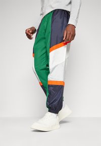 Groene, marineblauwe, witte en oranje trainingsbroek gemaakt van lichtgewicht, glanzend materiaal, met elastische boorden en een trekkoord in de taille.