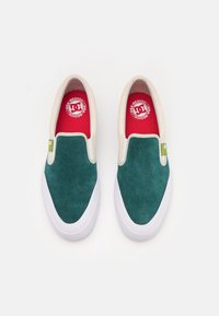 Zelené semišové slip-on boty s bílými gumovými podrážkami, s červenými stélkami a detailem zeleného loga, hladký povrch a kulatý tvar špičky.