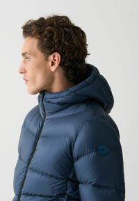 Junger Mann mit lockigem Haar, der eine marineblaue Steppjacke mit Kapuze trägt, schaut nach links vor einem schlichten, hellen Hintergrund.