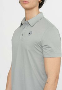 Polo shirt grigio chiaro realizzato in tessuto liscio, caratterizzato da un collo con tre bottoni e un piccolo logo in bianco e nero sul petto.
