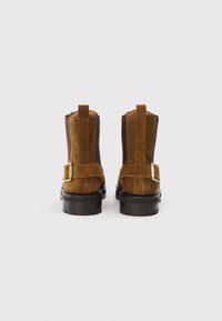Botine din suede maro, cu părți laterale elastice negre, cu bucle pentru tragere, talpă groasă neagră și catarame pătrate aurii în spate, prezentate dintr-o vedere din spate.