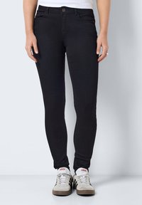 Black denim jeans with a skinny fit, featuring a classic button closure and front pockets. Worn with white and gray sneakers.