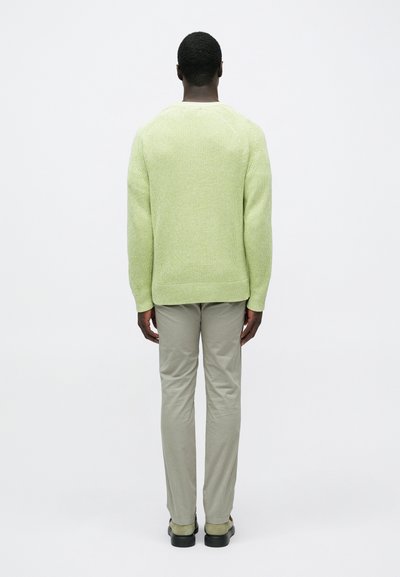 Muž stojaci chrbtom, oblečený v svetlozelenom pletenom svetri, béžových nohaviciach a zelených semišových topánkach na jednoduchom pozadí.