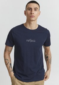 Jūrų mėlynos medvilnės marškinėliai su centriniu balto teksto grafiku "reflect." Trumpomis rankovėmis ir apvaliu iškirpimu. Lygi tekstūra.