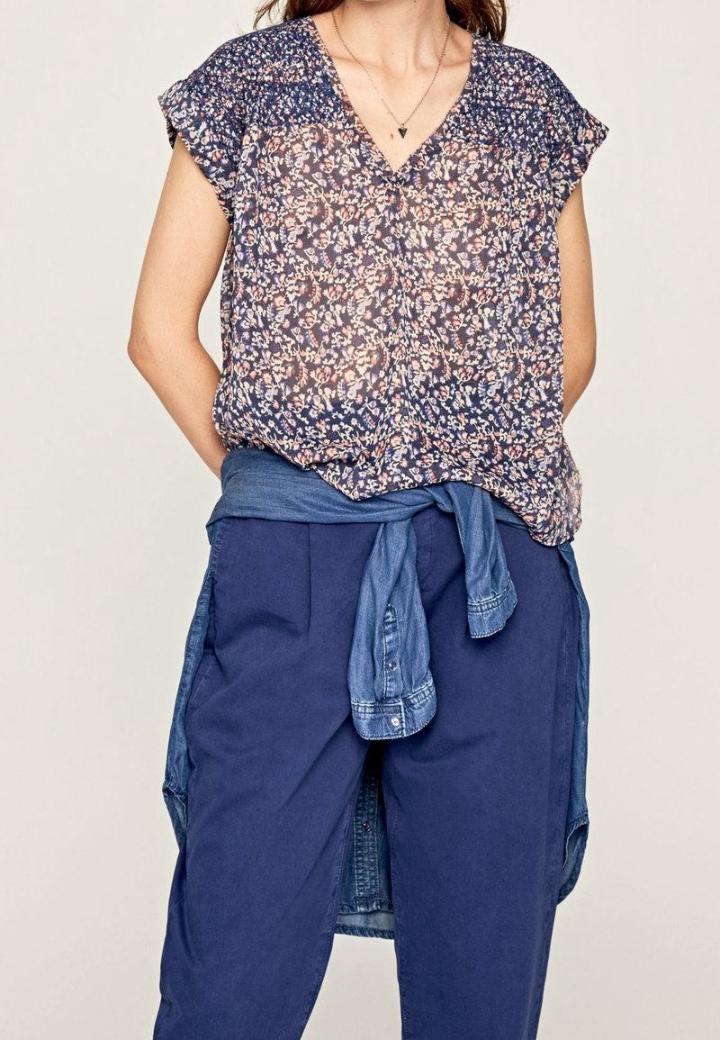 Blouse florale bleue marine avec des motifs rose clair et blancs, à manches courtes et décolleté en V. Portée sur un pantalon ample bleu foncé avec une chemise en denim nouée à la taille.