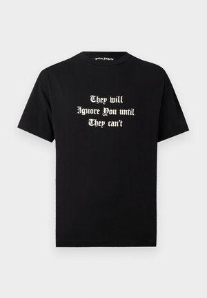 Jeune homme portant un T-shirt noir avec un texte blanc « They will Ignore You until They can’t » et un short noir avec des bandes latérales.