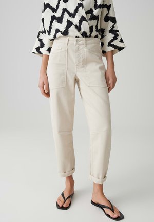 Frau trägt beige, hochgekrempelte Hosen mit großen Vordertaschen, eine Bluse mit schwarz-weißem Zickzackmuster und schwarze Flip-Flop-Sandalen.