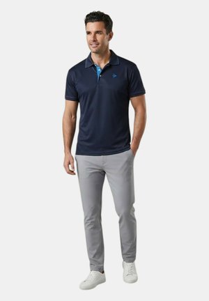 Uomo in piedi che indossa una polo blu navy, pantaloni grigio chiaro e scarpe da ginnastica bianche con una mano in tasca davanti a uno sfondo semplice.