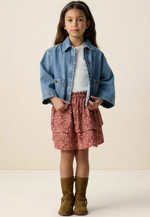 Jong meisje staand, gekleed in een blauwe denimjas, wit shirt, rode rokje met bloemen, bruine laarzen en een hoofdband.