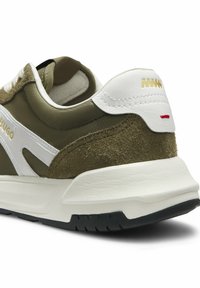 Gros plan sur le talon d'une basket vert olive et blanc avec le logo doré "Hugo", semelle blanche et matériaux en suède et cuir texturés.