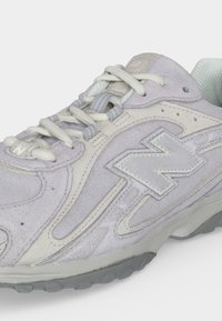 Ανοιχτό γκρι σουέτ αθλητικό παπούτσι με λευκά κορδόνια, μαλακό κολάρο και μεγάλο ραμμένο λογότυπο "N" στο πλάι, με γκρι ελαστική σόλα.