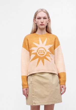 Jeune femme aux longs cheveux blonds portant un pull jaune et beige à motif soleil et une jupe beige mi-longue, debout sur fond blanc.