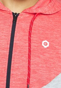 Röd zip-up hoodie med texturerad tyg, kontrasterande marinblå dragkedja, vitt logodetalj och justerbar röd dragsko med vita accenter.